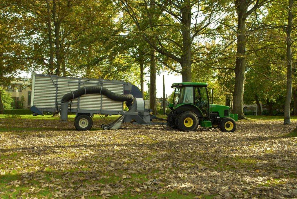 Ein moderner grüner Traktor mit Laubanhänger von Reinigungskonzepte Sachsen ist auf der Grünfläche eines Kunden im Einsatz. Es ist Herbst und die abgefallene Blätter bedecken die Umgebung. Unsere erfahrenen Mitarbeiter sorgen dafür, dass Ihre Grünflächen und Wege stets in einwandfreiem Zustand sind!