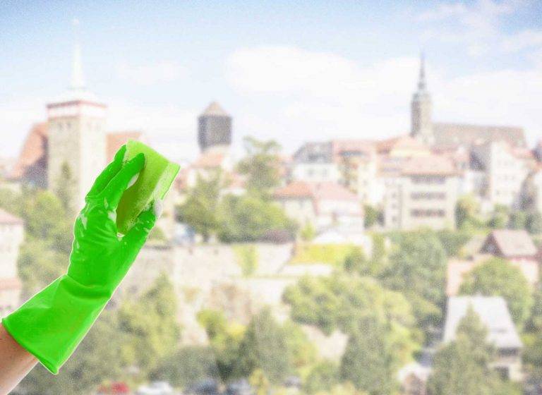Ein Reinigungskonzepte-Mitarbeiter mit Handschuh reinigt ein Fenster, die Stadt Bautzen ist im Hintergrund sichtbar.