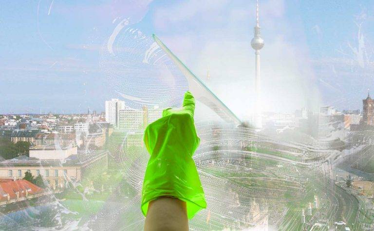 Ein Reinigungskonzepte-Mitarbeiter reinigt mit einem Glasabzieher ein Fenster, im Hintergrund ist Berlin erkennbar.