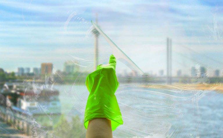 Ein Reinigungskonzepte Mitarbeiter putzt eine Fensterscheibe. Hinter dem Glas kann man unscharf Düsseldorf sehen.